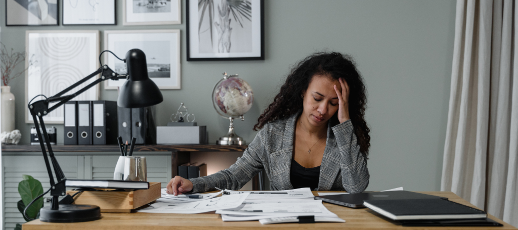A woman sits at a cluttered desk, holding her head in frustration amid papers, a laptop, and stationery. A lamp illuminates her struggle as she contemplates psychosocial risk controls. Behind her, a globe rests on the shelf while framed pictures adorn the wall with silent reassurance.