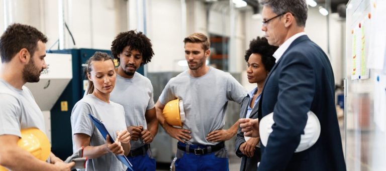 A group of factory workers in gray shirts and a manager in a suit gather for a discussion in an industrial setting. Some workers hold yellow safety helmets, and one person writes on a clipboard.