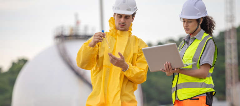 Two engineers wearing safety gear, including helmets and vests, work outdoors at an industrial site; one holds a test tube while the other uses a laptop, with large storage tanks in the background.