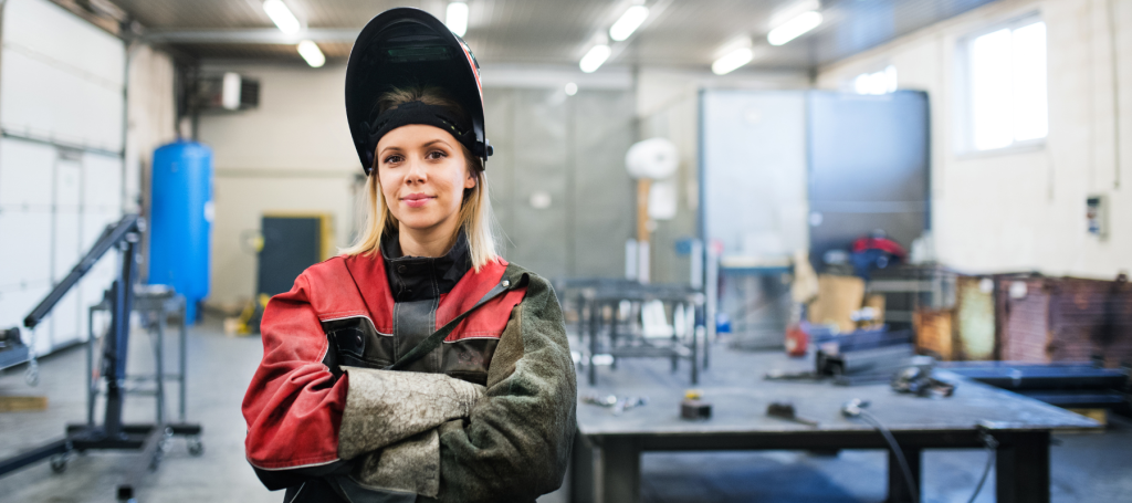 A woman wearing welding gear and a helmet stands confidently with crossed arms in a workshop, surrounded by industrial equipment and metalwork tables.